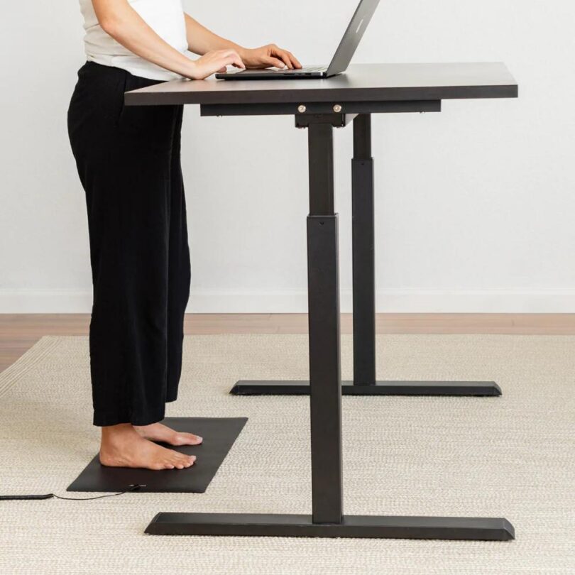 A woman stands barefoot on a black grounding mat while working at a standing desk with a laptop